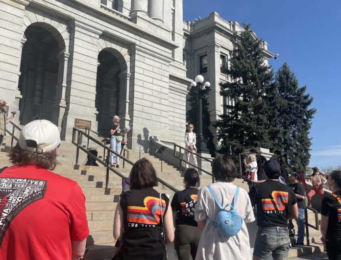 A group of people in colorful t-shirts watch a woman with short blonde hair in jeans speak on the steps of the Colorado capitol.