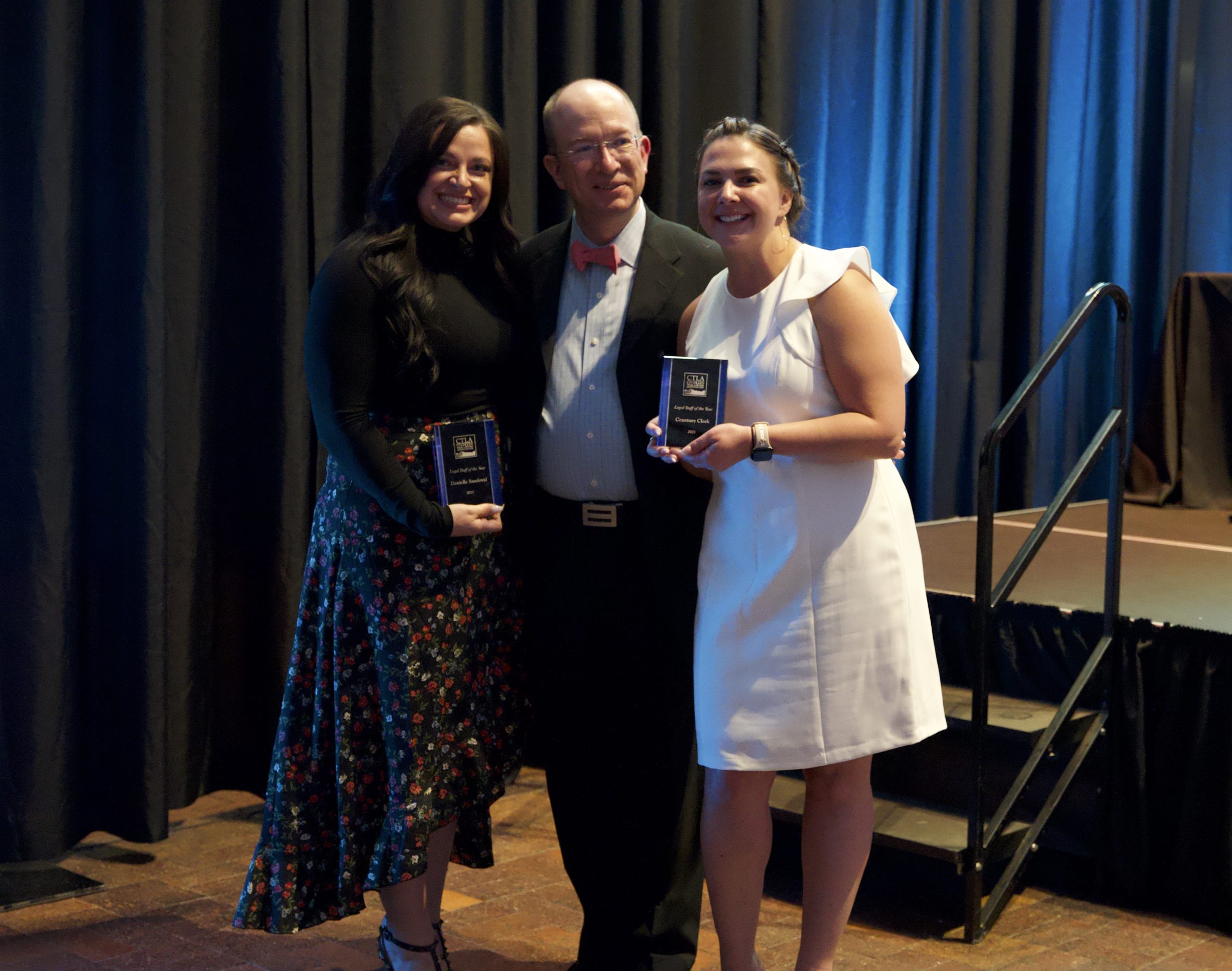 Three people smile and pose for the camera close together. On the left, a woman with dark hair in a black shirt and patterned skirt smiles and holds a plaque. In the middle, a middle aged bald man in a suit with a red bowtie smiles slightly. On the right, a blonde woman in a white dress smiles and holds a plaque.