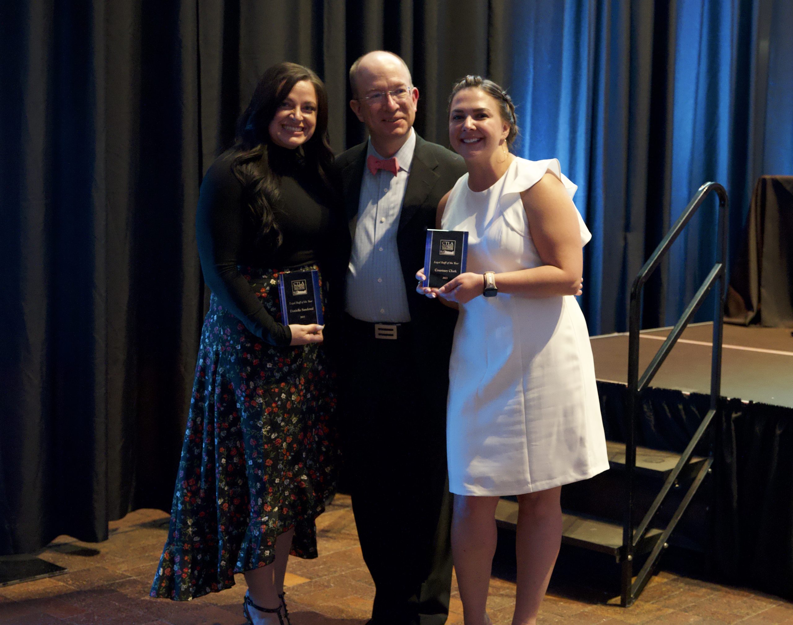 Three people smile and pose for the camera close together. On the left, a woman with dark hair in a black shirt and patterned skirt smiles and holds a plaque. In the middle, a middle aged bald man in a suit with a red bowtie smiles slightly. On the right, a blonde woman in a white dress smiles and holds a plaque. 