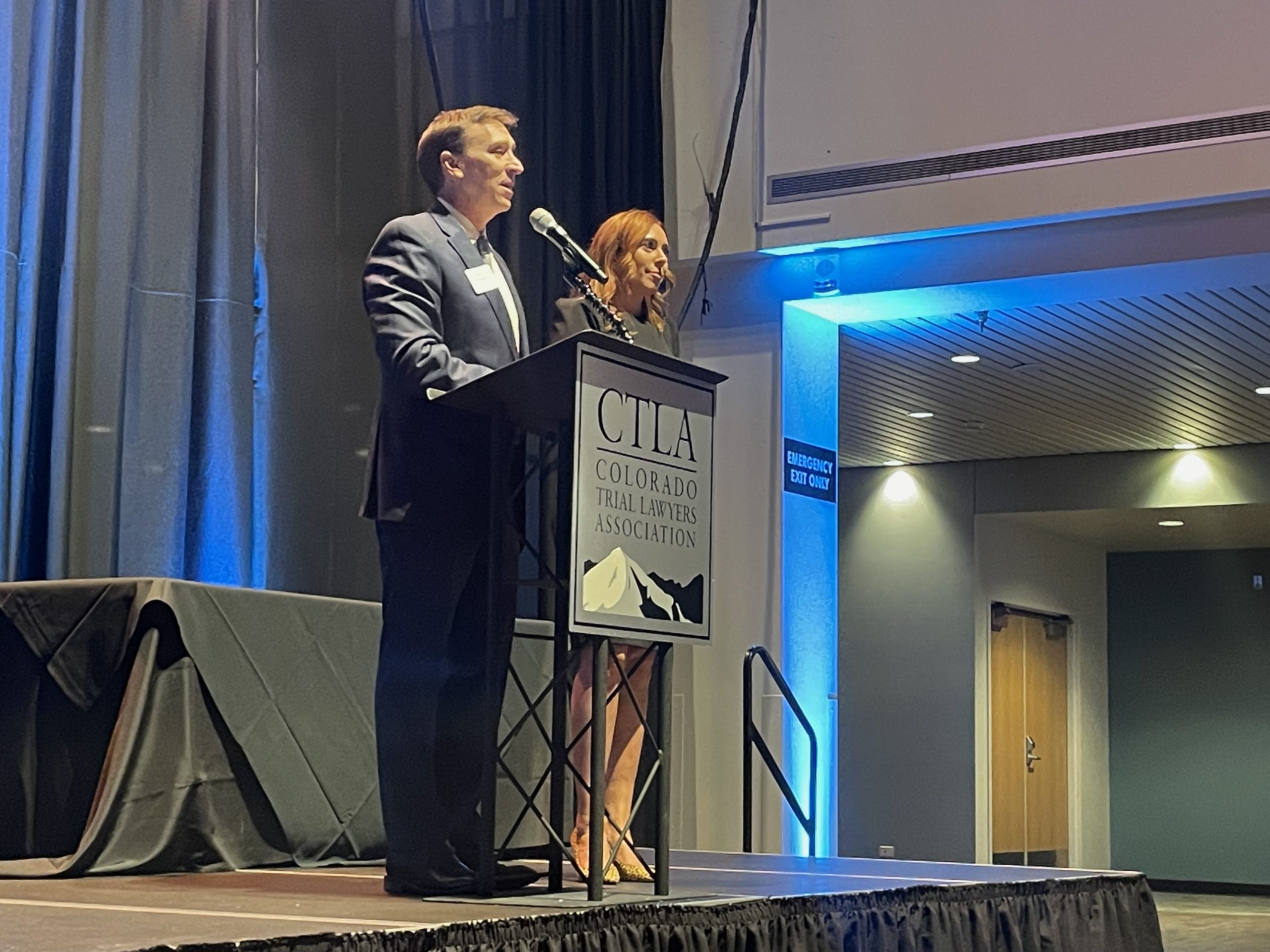A middle aged man in a dark blue suit stands and speaks at a podium with a sign that reads CTLA Colorado Trial Lawyers Association. To his right a woman with red hair in a black dress stands and smiles slightly.