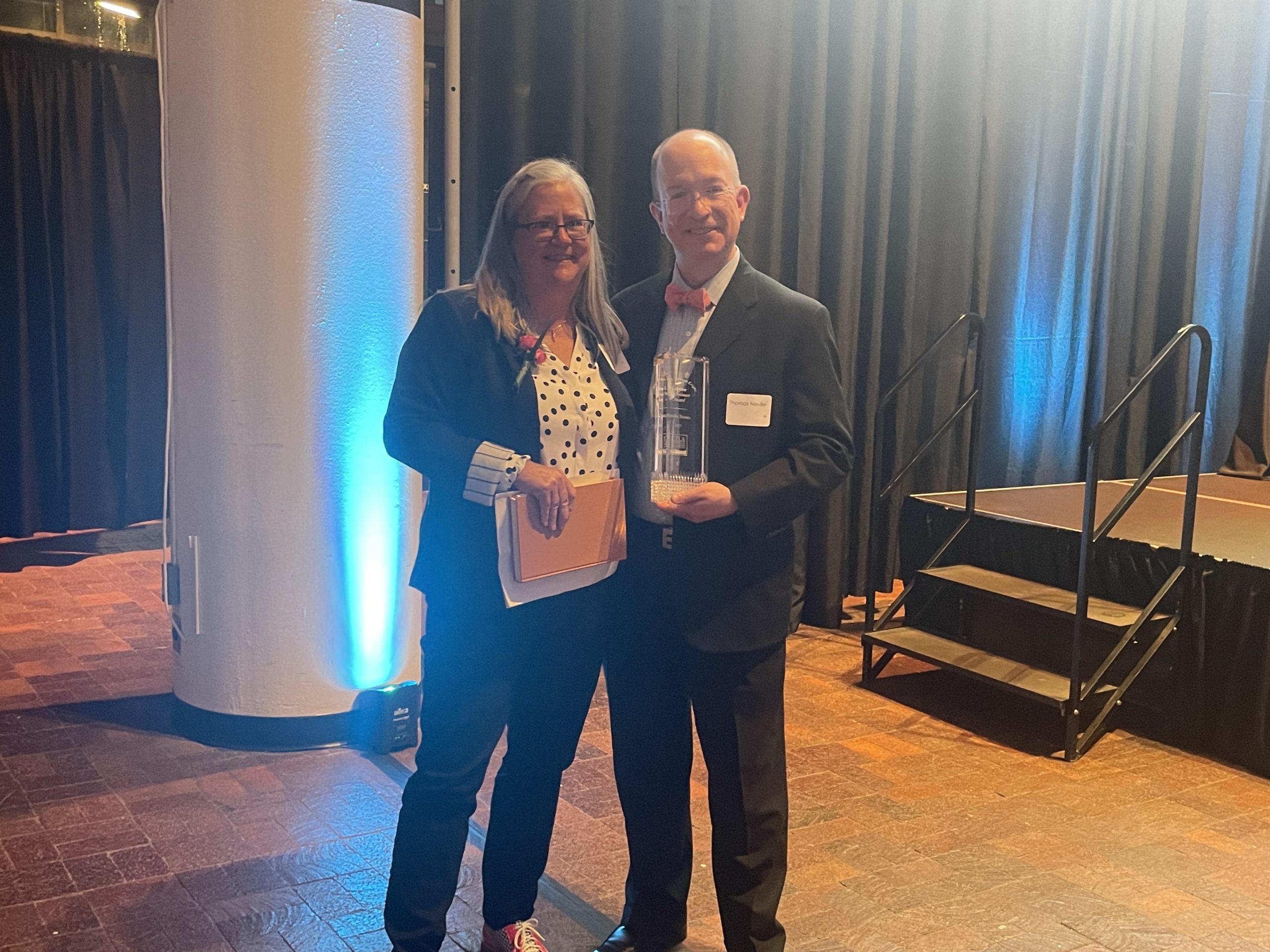 A middle aged woman with gray hair wearing a black pantsuit with a polka dot shirt underneath holding a manilla folder and papers smiles and poses alongside a middle aged bald man in a suit with a red bowtie holding a crystal award. 