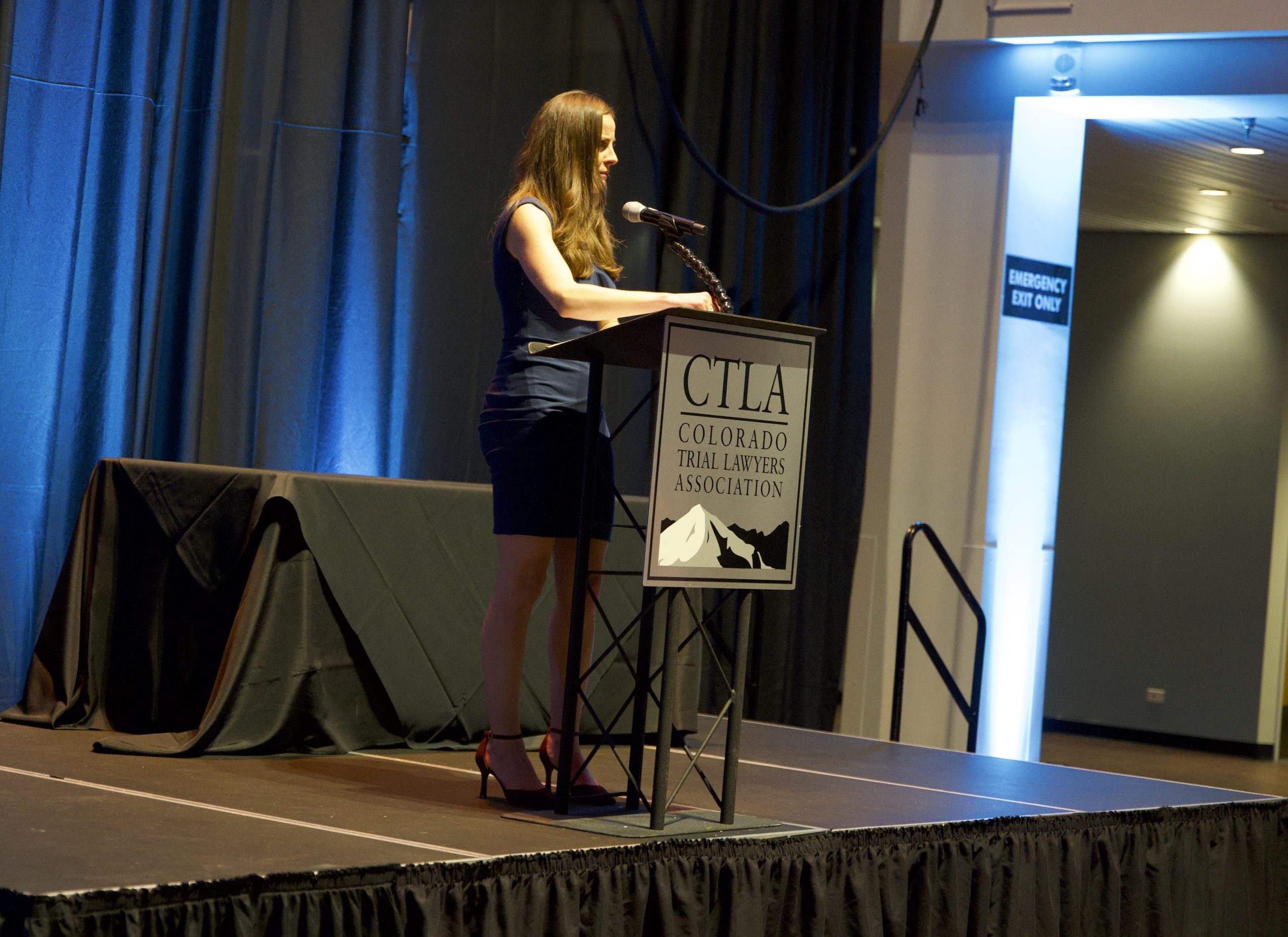 A young woman with long brown hair wearing a dark blue dress and heels stands in front of a podium on a stage that reads CTLA Colorado Trial Lawyers Association.