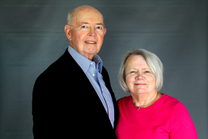 A man wearing a suit coat and a woman wearing a pink top pose for a picture with a gray background.