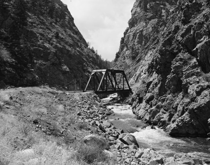 a black and white photo shows a bridge overlooking a river with steep looking mountains on either side