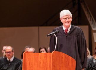 Commission on Judicial Discipline Recommends Censure of Former Chief Justice Nathan Coats A man with white hair, round glasses in a judge's robe smiles in front of a speaker's podium in front of a crowd.