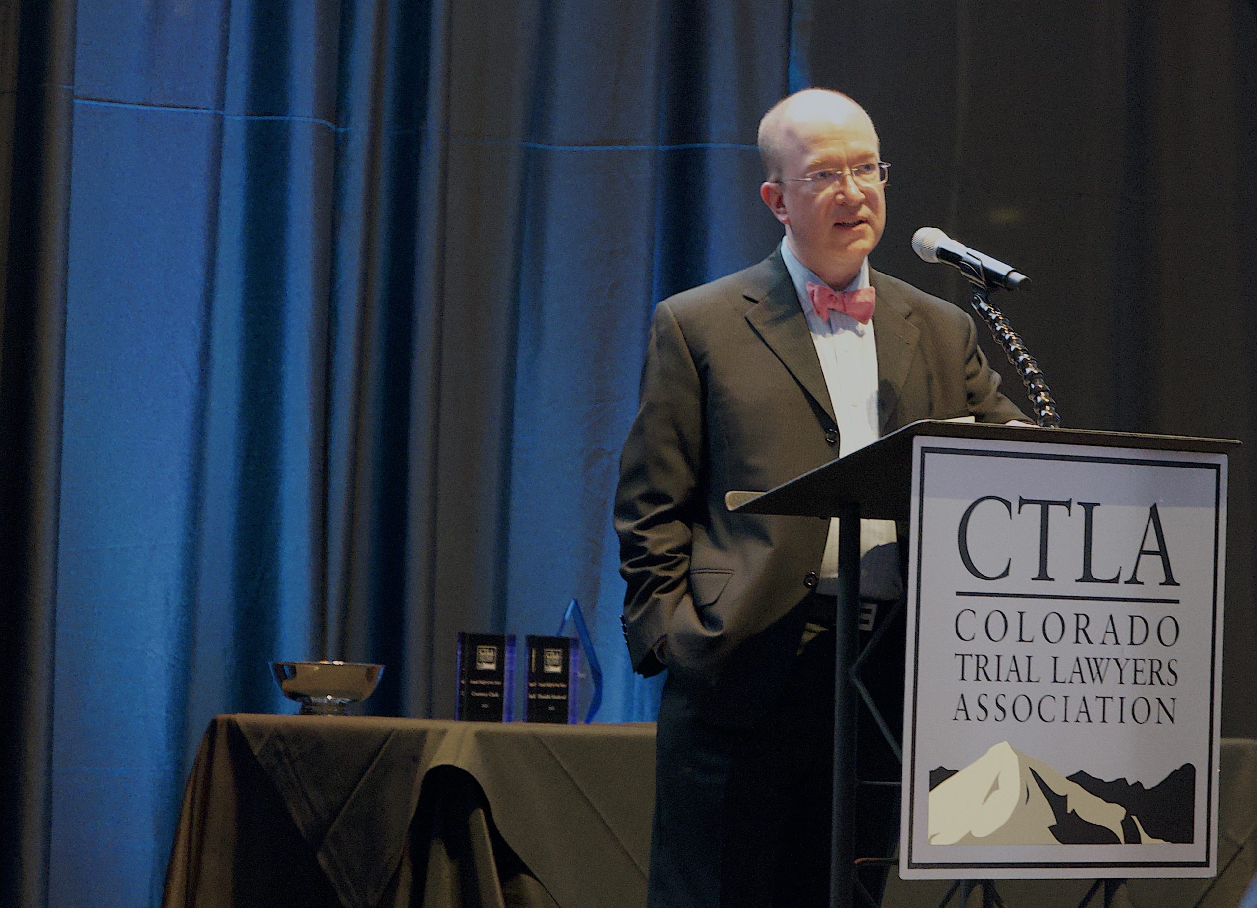 A middle aged bald man in a suit with a red bowtie speaks at a podium on a stage that reads CTLA Colorado Trial Lawyers Association.