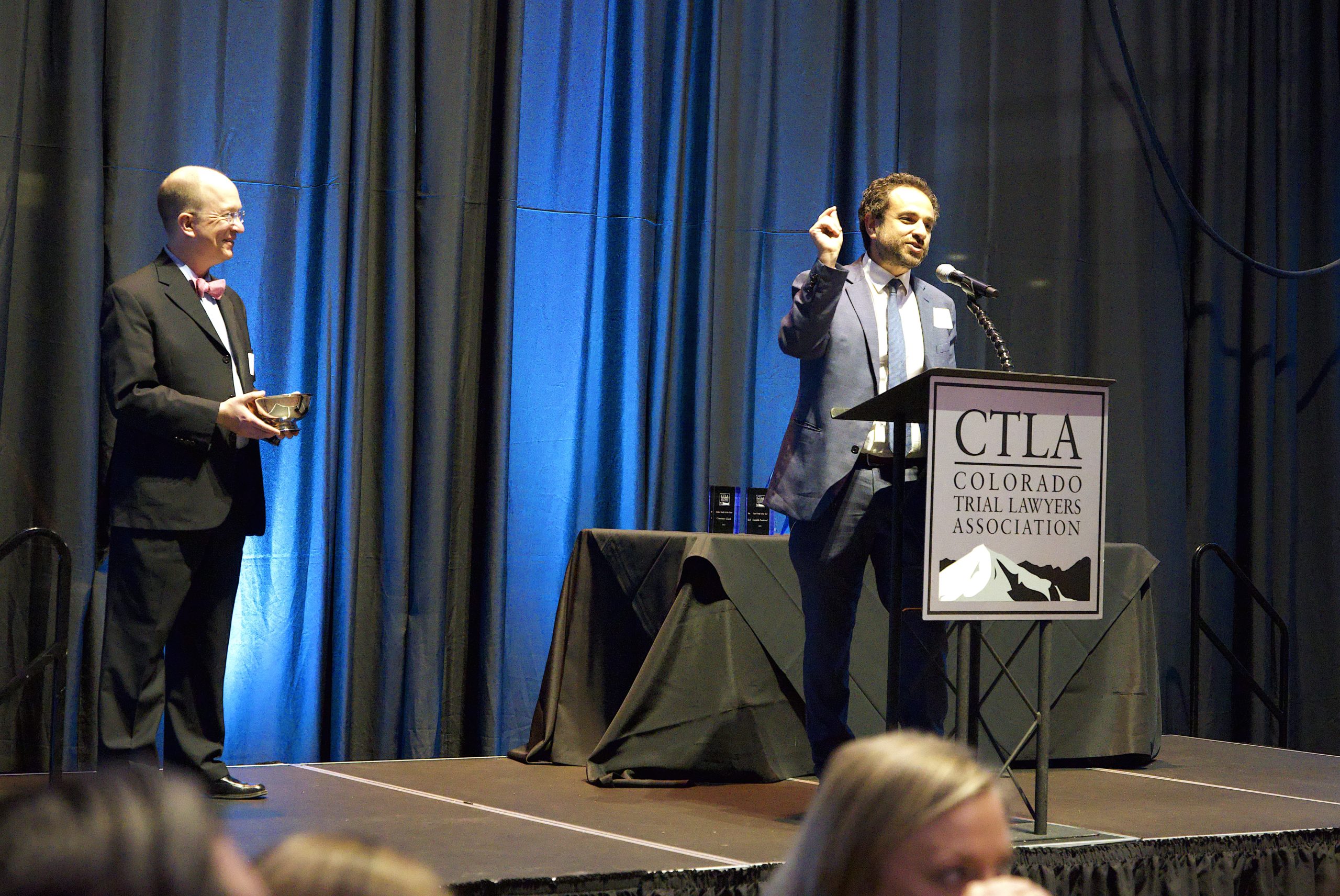 A man with brown hair in a dark suit stands in front of a podium on a stage with a sign that reads CTLA Colorado Trial Lawyers Association. One hand is raised in emphasis as he speaks. To his right, a middle aged bald man in a suit with a red bowtie holds a silver bowl award. 