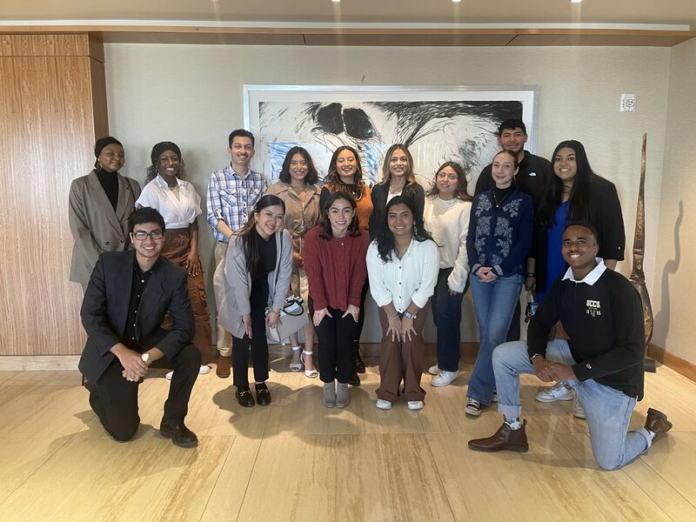 A group of people from many different backgrounds pose for a picture with a unique image or painting behind them.