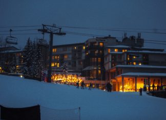A Decade of Legal Stress Before Long-Planned Burnt Mountain Ski Expansion Snowmass Village at night. A ski lift is backlit with soft, glowing lights from a ski resort. People are walking around in the foreground and a fresh layer of powdery snow coats the ground and nearby trees.