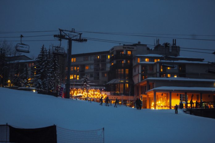 Snowmass Village at night. A ski lift is backlit with soft, glowing lights from a ski resort. People are walking around in the foreground and a fresh layer of powdery snow coats the ground and nearby trees.