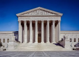 U.S. Supreme Court Keeps Indian Child Welfare Act in Place A white building with seven pillars in front with a peaked concrete roof has steps coming up to it. Statues of a person sitting in a chair are seen on both arms of the stairs.
