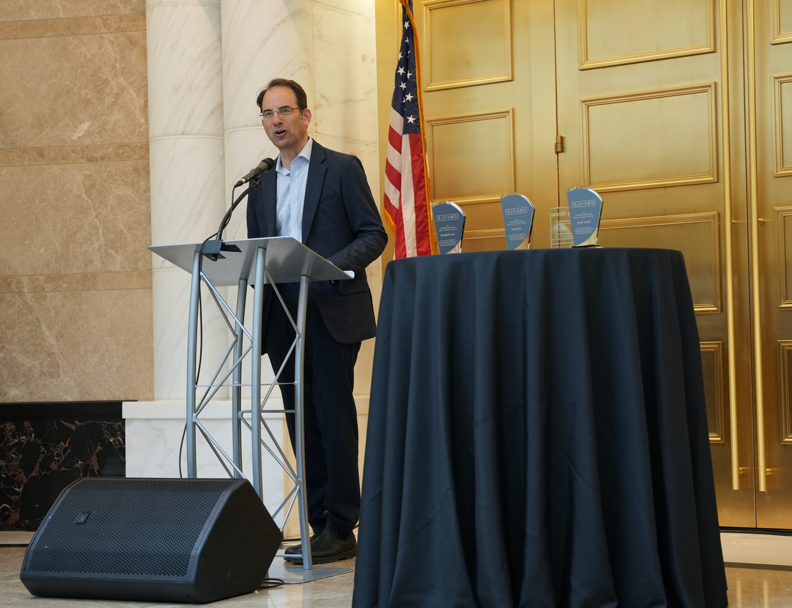 A middle aged man with dark hair and glasses wearing a dark suit stands in front of a podium with a microphone and speaks. In front on him is a table with three awards, behind him is a marble wall, gold doors and an American flag. 