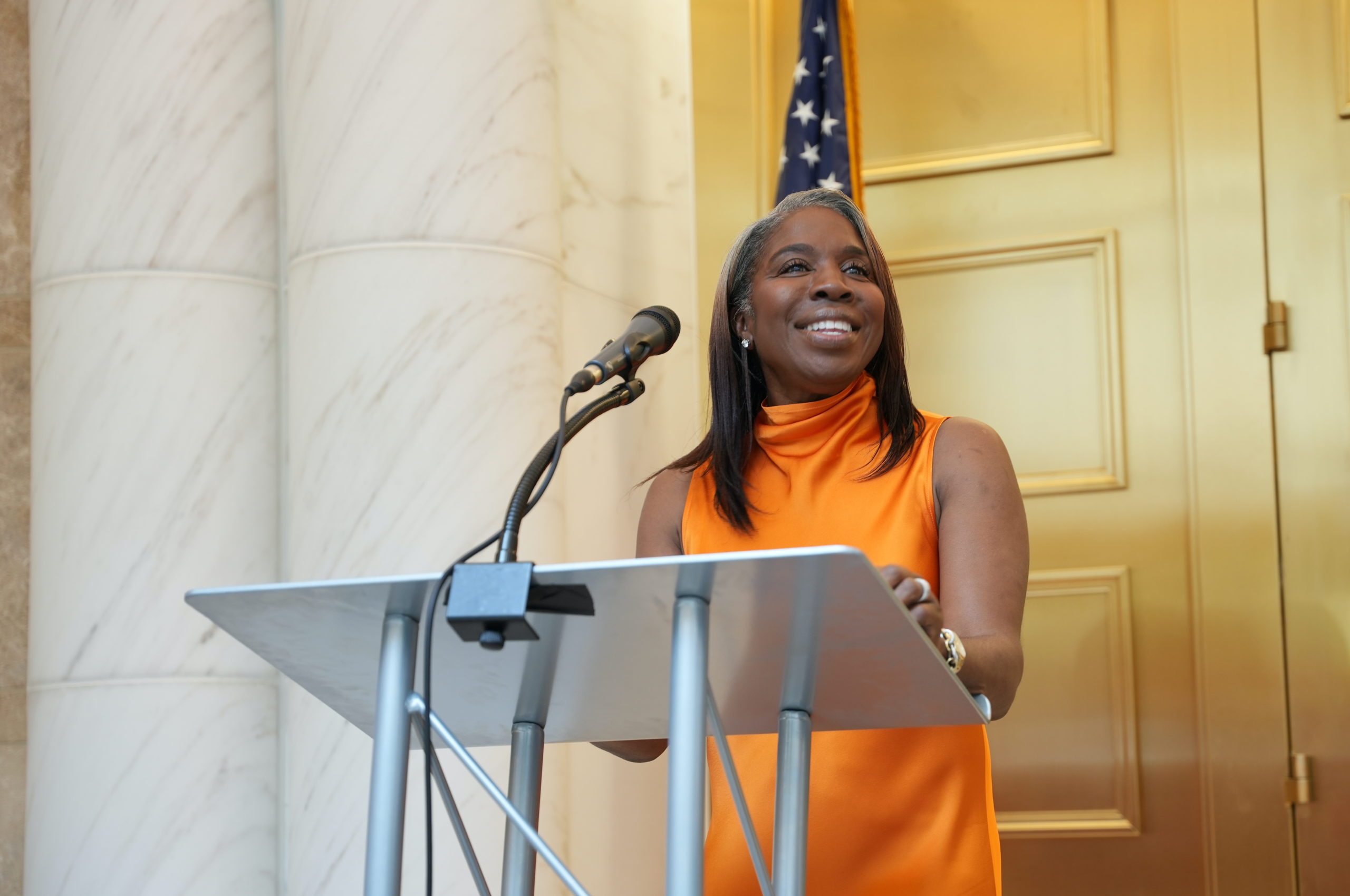 A middle aged Black woman in an orange formal dress stands in front of a podium with a microphone and smiles. In the background are gold doors, marble pillars and an American flag.