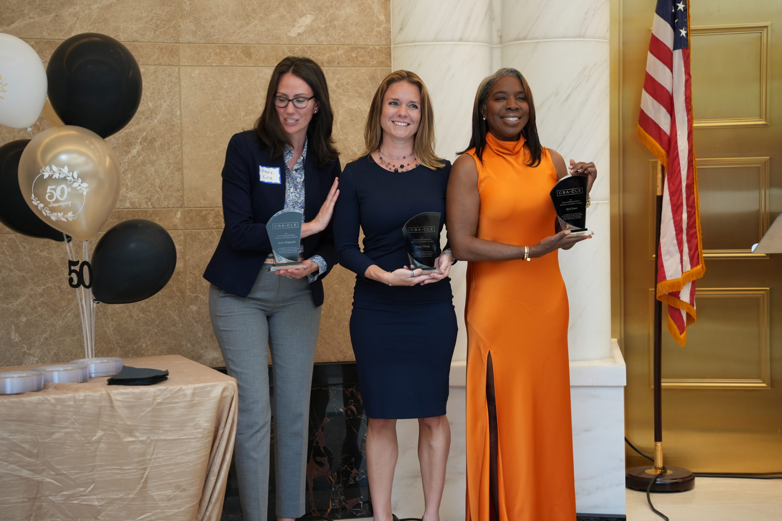 Three women in formal clothes stand in a line and smile while holding dark stone awards in front of marble walls and white pillars. 