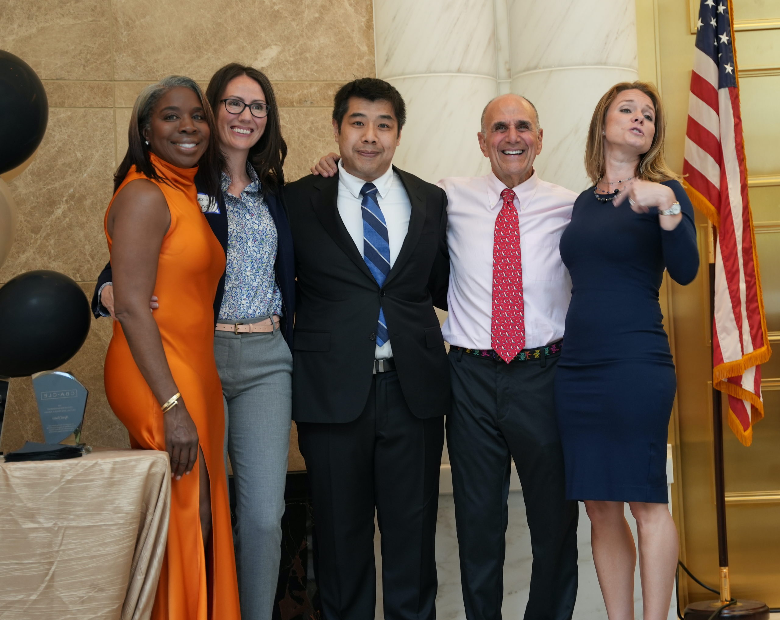 Five people in formal clothes stand in a line and smile in front of white pillars, marble walls, gold doors and an American flag. 