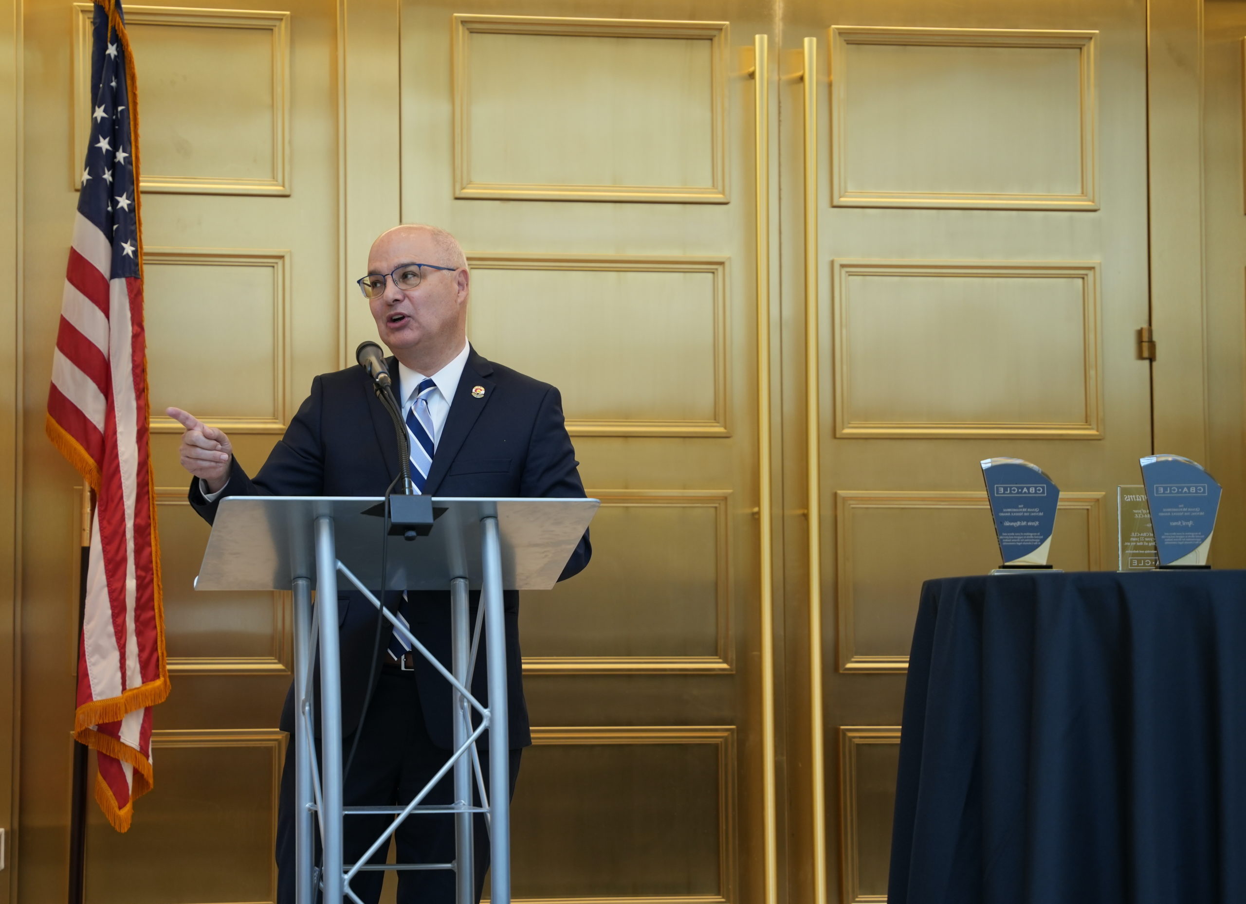 A middle aged man in a dark suit wearing glasses stands in front of a podium with a microphone and points to the left while speaking. To the right is a table with three awards. In the background are gold doors and an American flag. 