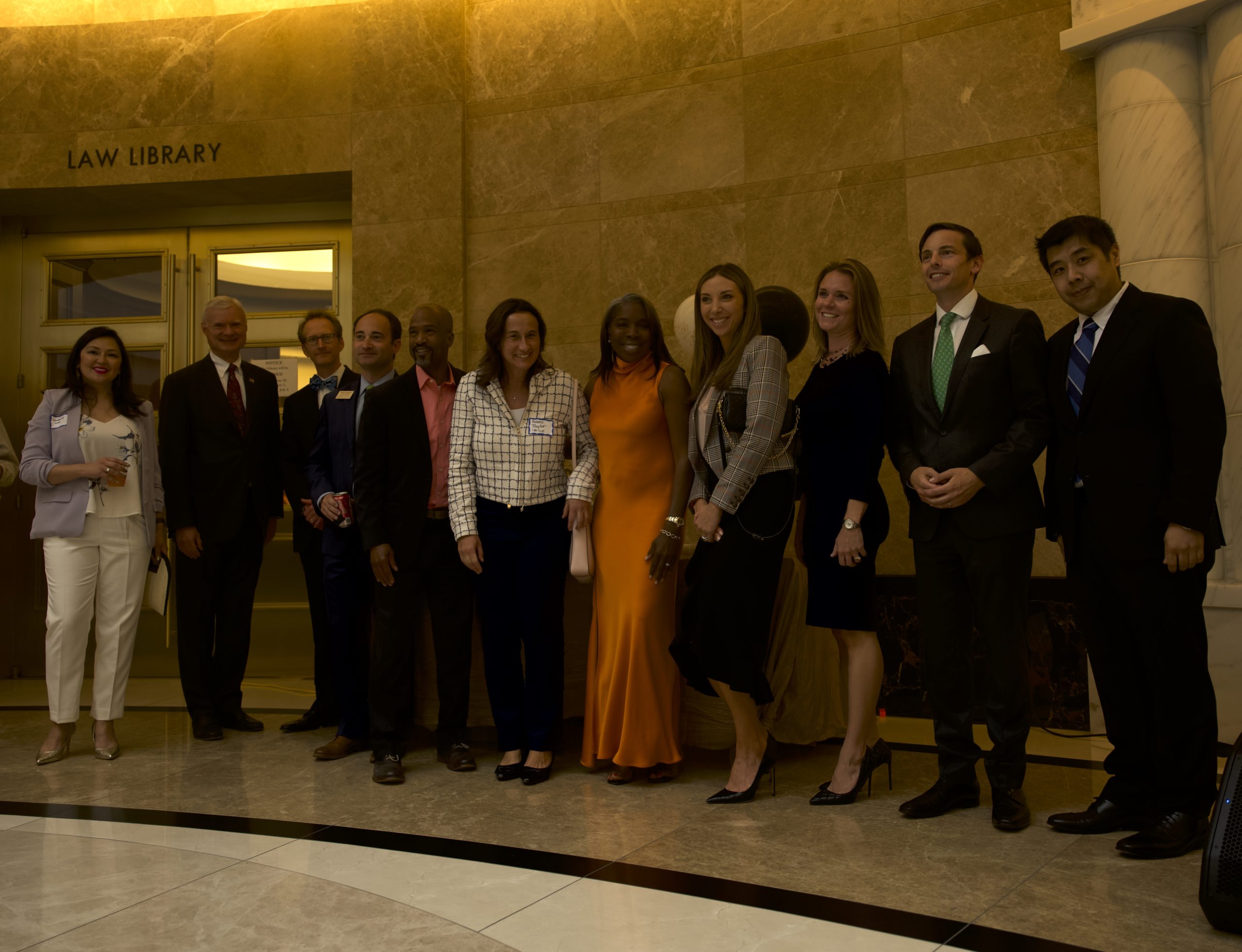 A group of people in formal wear stand in a line and smile in front of a rounded marble wall.