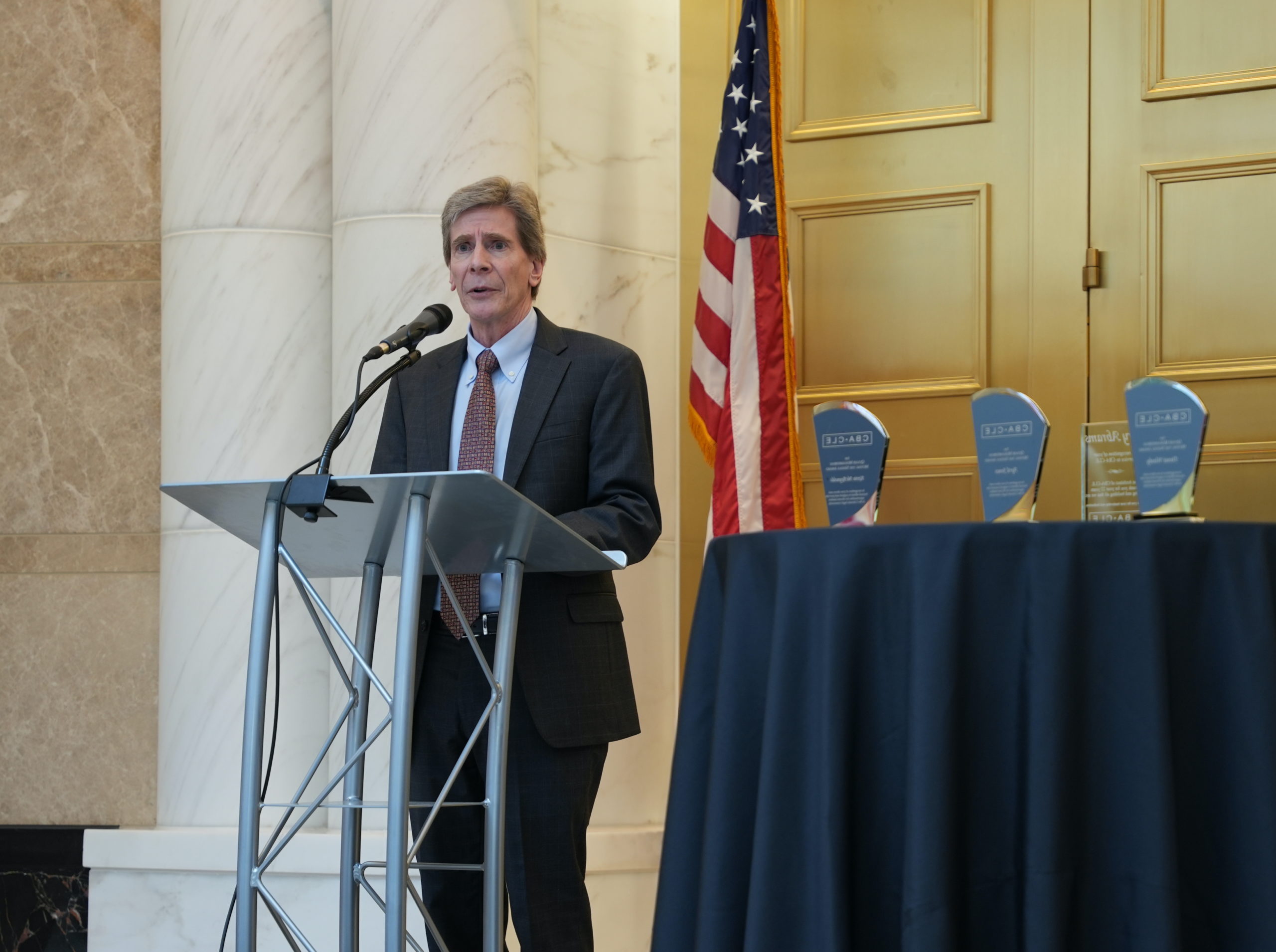 An older man with gray hair in a dark suit stands at a podium with a microphone and speaks. In front on him is a table with three awards, behind him is a marble wall, gold doors and an American flag.