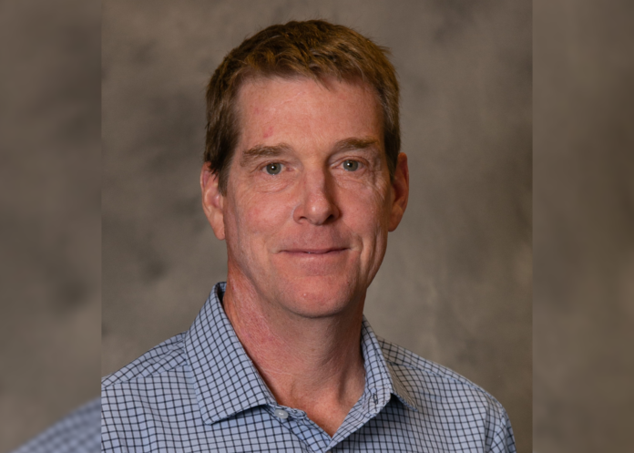 man poses for a photo with a gray background. He’s wearing a blue-checkered shirt.