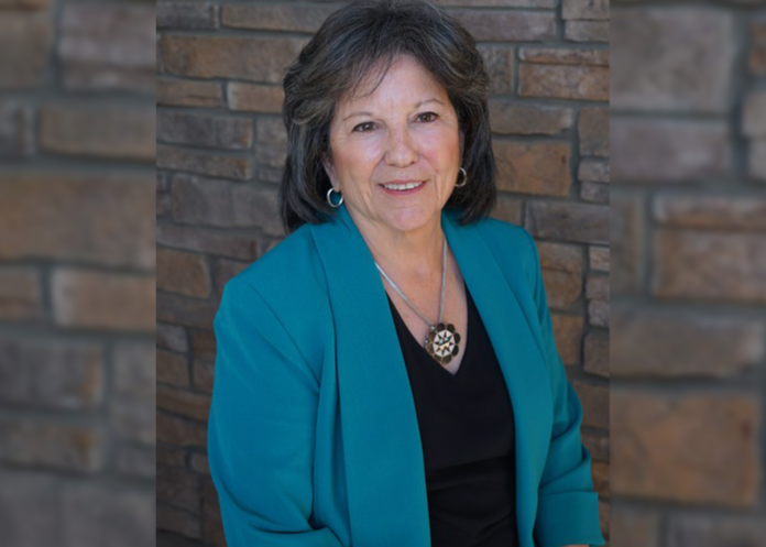 A woman in a blue blazer and black blouse is smiling warmly in front of a brick wall.