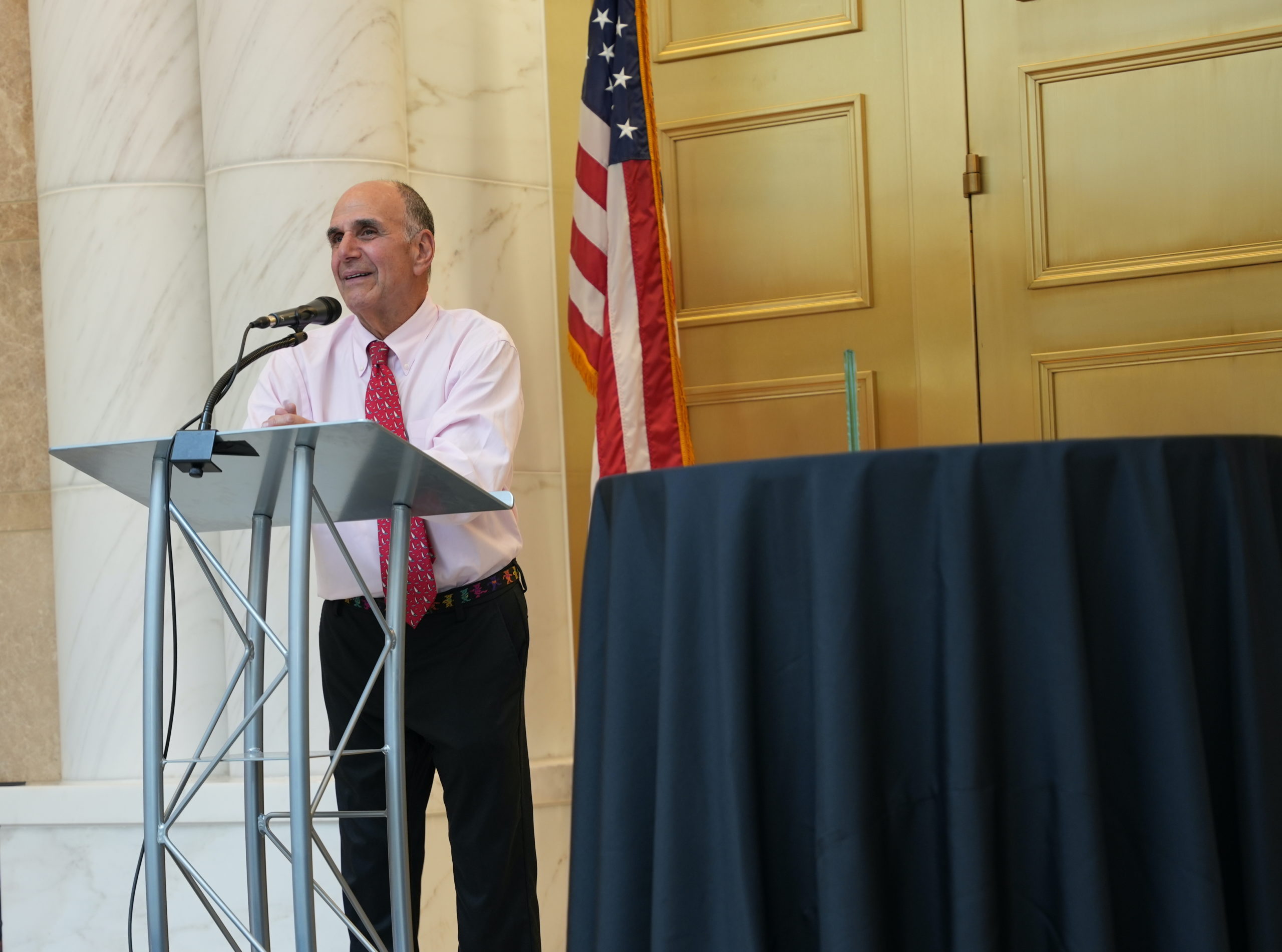 An older man in a white button up shirt and red tie stands in front of a podium with a microphone and smiles slightly while speaking. In the foreground is a table with a glass award. In the background are gold doors, marble pillars and an American flag. 