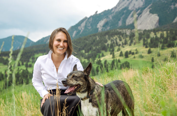 A woman kneels as she poses with a dog. Trees and part of a mountain can be seen in the background.
