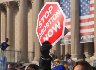 Suit Hopes to Overturn Colorado Law Against ‘Sidewalk Counseling’ Outside Abortion Clinics A person holds a sign that looks like a red stop sign with the text “Stop Abortion Now” above a crowd of people in front of a government building.