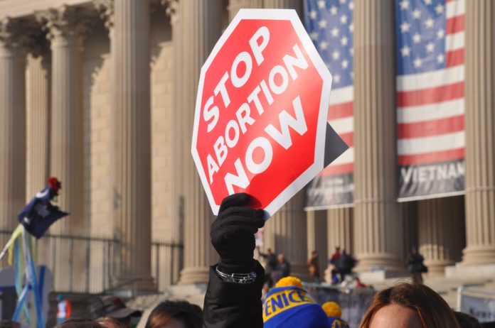 A person holds a sign that looks like a red stop sign with the text “Stop Abortion Now” above a crowd of people in front of a government building.