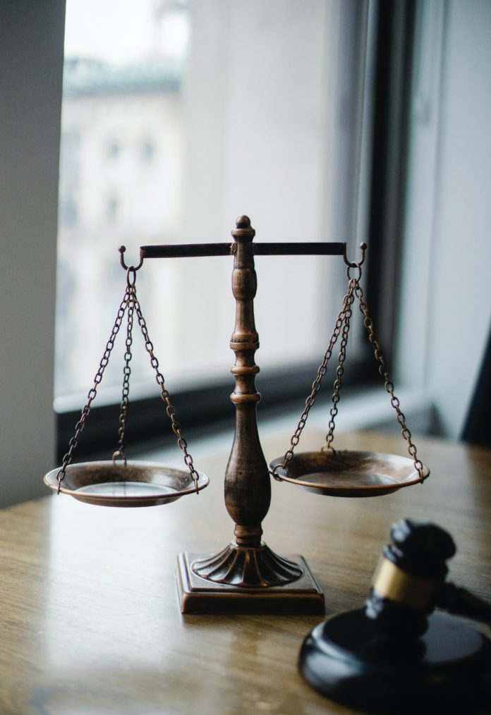 Two scales are attached by chains to upper hooks that are supported by a small column. It is the “scales of justice.” In the foreground a gavel can be seen.