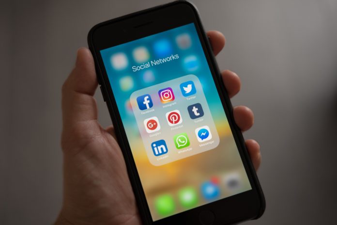 A hand holds an iPhone open to a folder titled ‘social networks.’ The folder has app icons for Facebook, Instagram, Twitter, Google, Pinterest, Tumblr, LinkedIn, Whatsapp and Facebook Messenger.