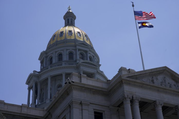 The Colorado Capitol Building. A close up image of an official building with a gold dome over a rotunda with pillars. On the right of the dome is a flag pole with the American flag above the Colorado flag.