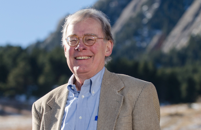 An older man with white hair and glasses wearing a checkered blazer and a blue button-up shirt smiles outside with the mountains in the background.