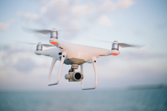 josh-sorenson-ouuigywbXlI-unsplash A small white machine with propellers attached to it flies in the air. What appears to be a camera is attached.