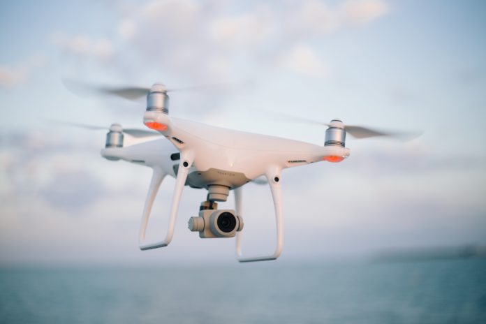 A small white machine with propellers attached to it flies in the air. What appears to be a camera is attached.