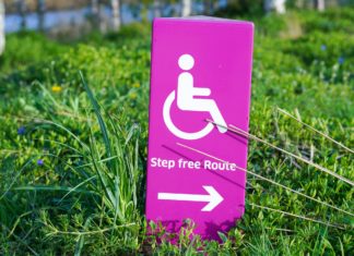 33rd Anniversary of ADA Commemorated A pink sign with a symbol of someone using a wheelchair. It says “Step free route” with an arrow pointing to the right below it. Grass is strewn about.