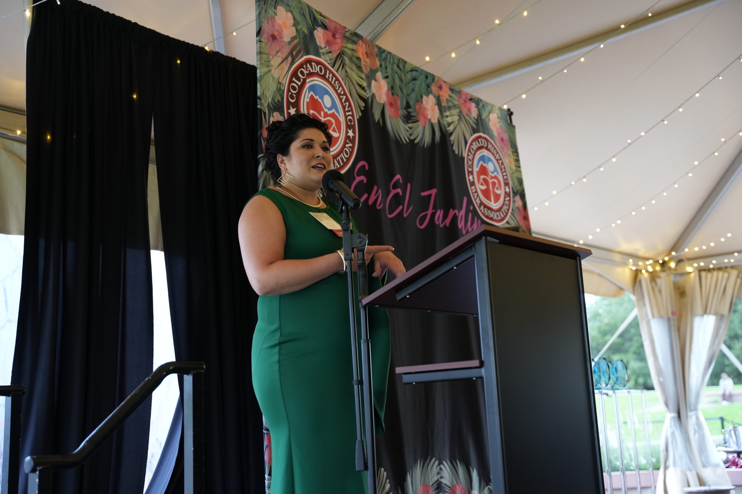 A younger Hispanic woman with black hair in a formal dark green dress with gold jewelry and hair in an updo speaks in front of a podium on a stage. Behind her is a banner that reads En el Jardín.