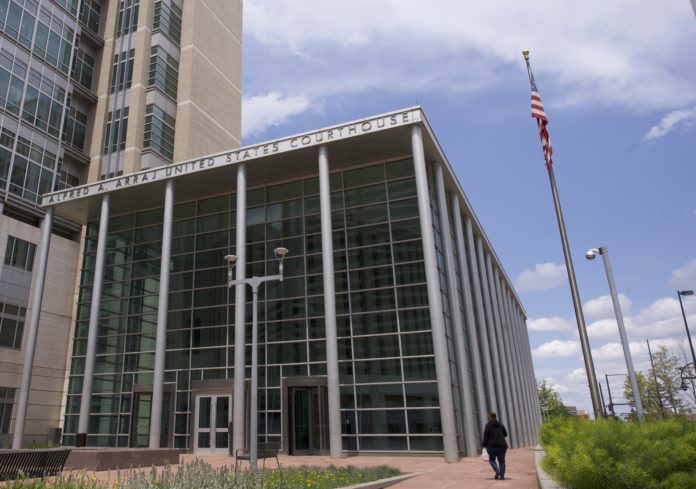 A modern looking courthouse with letters on the top that read “Alfred A. Arraj United States Courthouse.” The courthouse has glass on the front of the building and six light gray pillars in front. In front of the courthouse is a small courtyard with benches and wildflowers. To the right of the courthouse a woman walks towards the door and there’s a flagpole with the American flag.