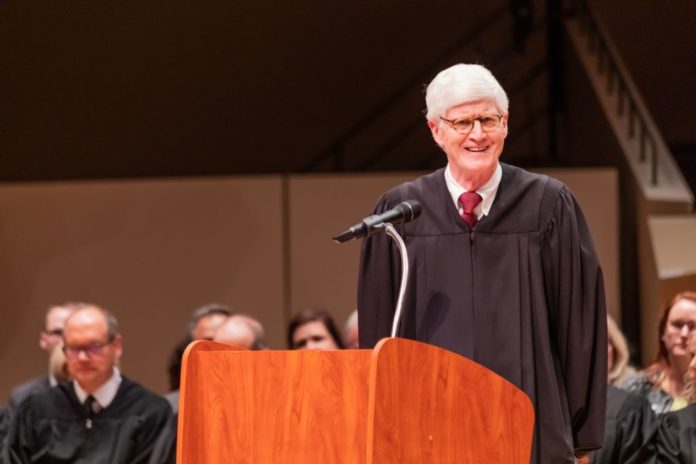 A man with white hair, wearing a judge's robe with a tie and glasses on, looks out in front of a podium with a microphone. Multiple people can be seen sitting in the background.