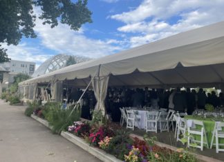 Colorado Hispanic Bar Association Banquet Spotlights Mission Progress and Challenges A white events tent with fold up tables and chairs inside and a crowd of people in formal clothes. On the outside of the tent are a row of colorful flowers and behind the tent is a glass domed greenhouse.