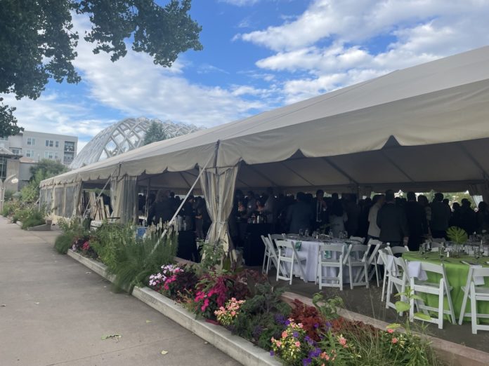 A white events tent with fold up tables and chairs inside and a crowd of people in formal clothes. On the outside of the tent are a row of colorful flowers and behind the tent is a glass domed greenhouse.