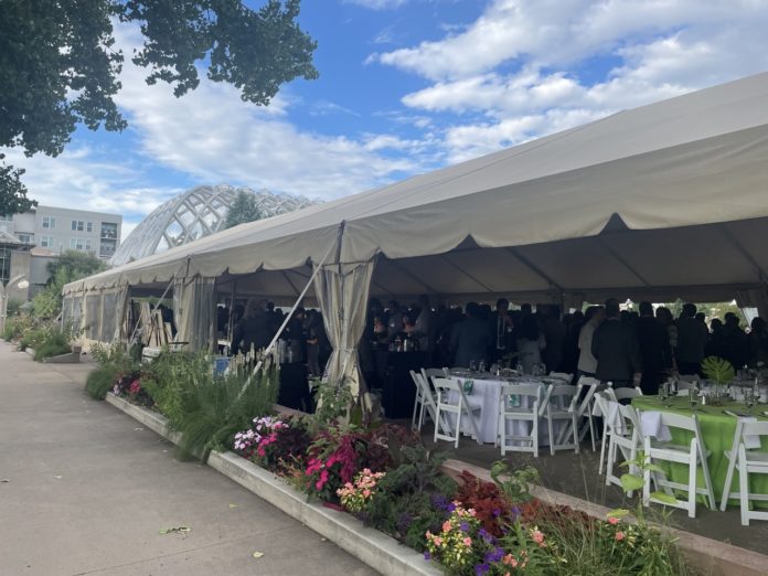 IMG_1047 copy Large A white events tent with fold up tables and chairs inside and a crowd of people in formal clothes. On the outside of the tent are a row of colorful flowers and behind the tent is a glass domed greenhouse.