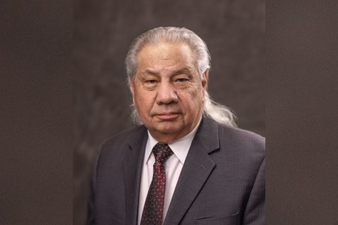 An older man with long gray hair behind his shoulders in a gray suit and red tie looks at the camera with a serious expression.