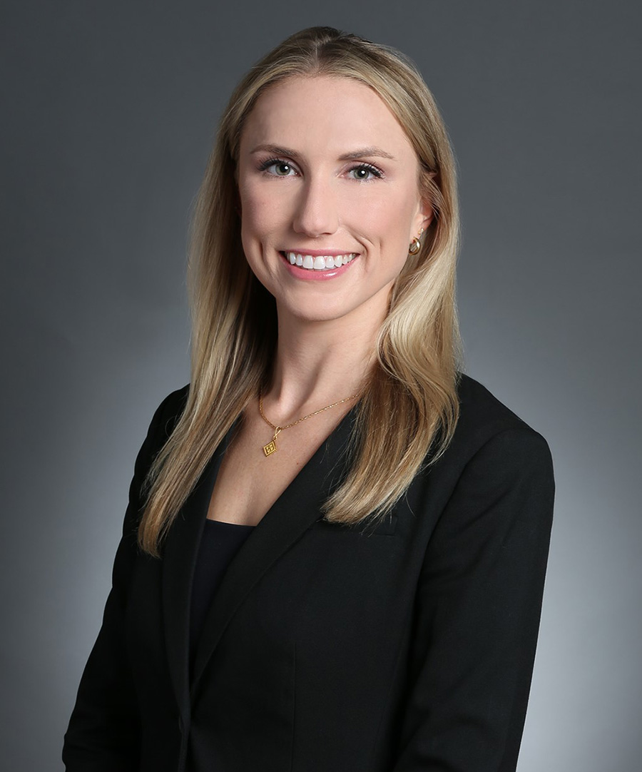 A younger blonde woman wearing a black blazer and gold jewelry smiles in front of a gray background. 