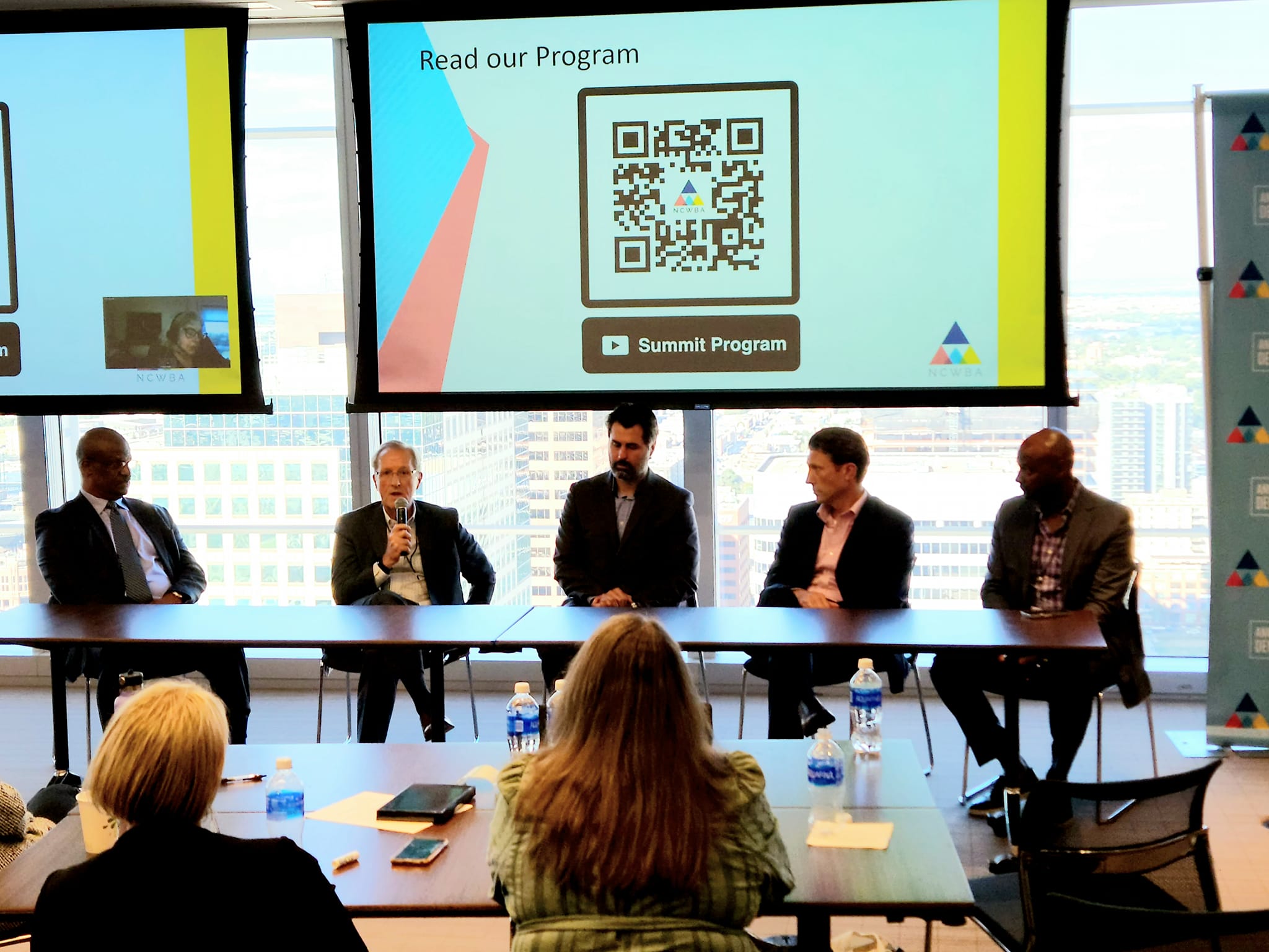 ony Holloway, Gene Commander, Chris Stanton, Jared Briant and C.J. Chapman sit at a table in front of a crowd. Behind the panelists is a presentation slide on a large screen in front of floor-to-ceiling windows.