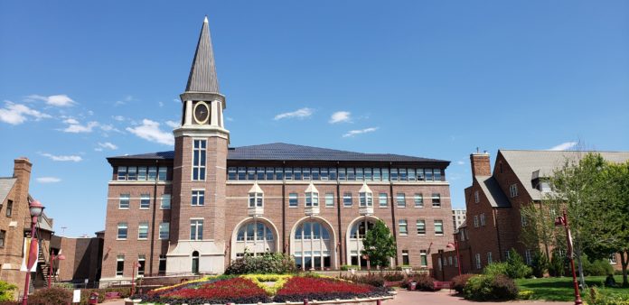 A large building with a pointed tower. In front of it are many flowers that are arranged. There are also buildings on the sides of the building in the center.