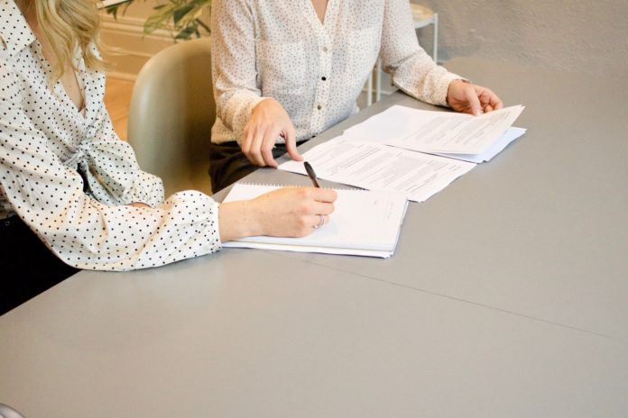 An office table with three stacks of papers on it. On one side of the table are two women in professional clothes. One woman gestures toward a stack of papers the other woman is signing with a pen.