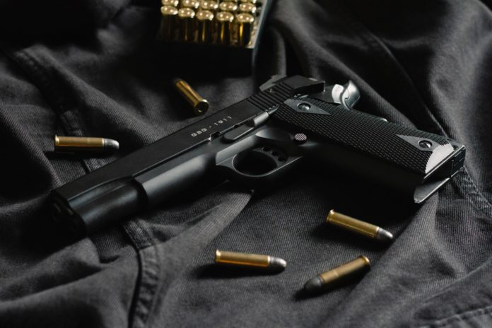 A black firearm has golden colored bullets surrounding it as it all lays on a black surface.