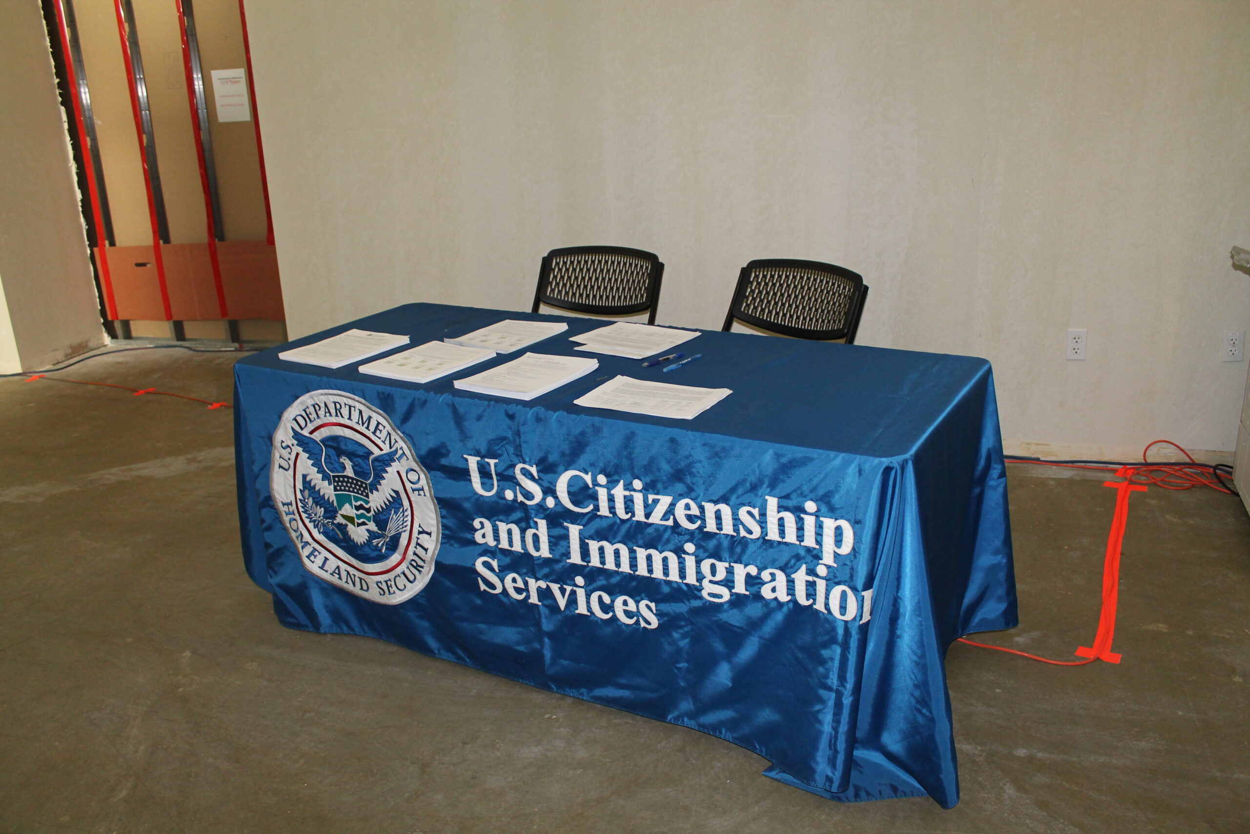 a table has forms stacked on top and two chairs behind it. There’s a blue satin tablecloth over it with a seal on the left and the text U.S. Citizenship and Immigration Services.