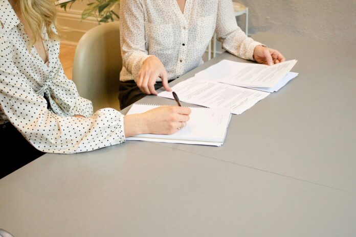 two people are reviewing and signing papers on a conference table while speaking to each other