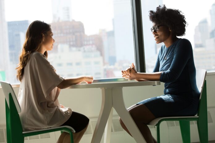 Two professional women sit at a table having a conversation.